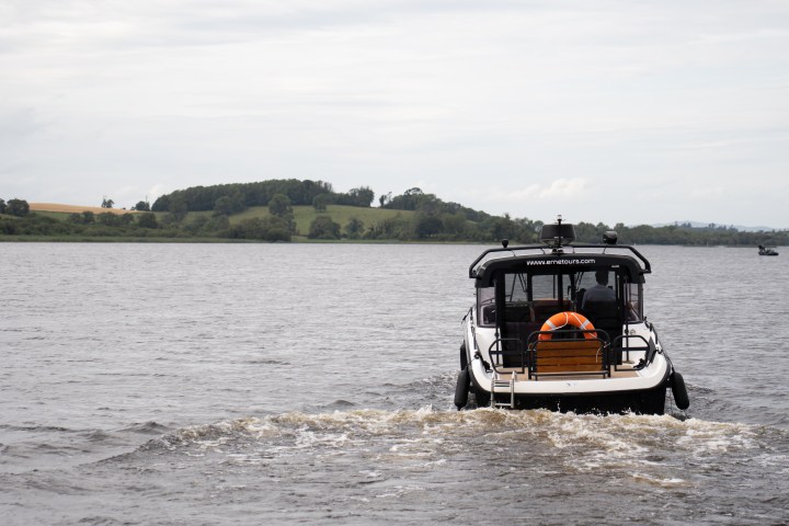 Boat with orange life ring on water, heading towards tree-covered hills under cloudy sky.