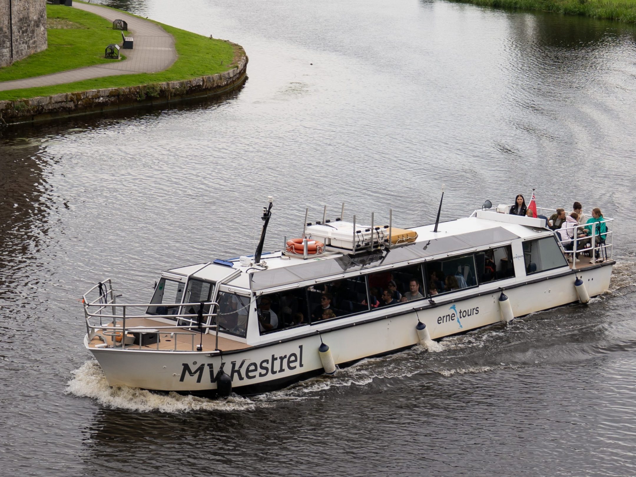 Tour boat on river near stone building and green landscape.