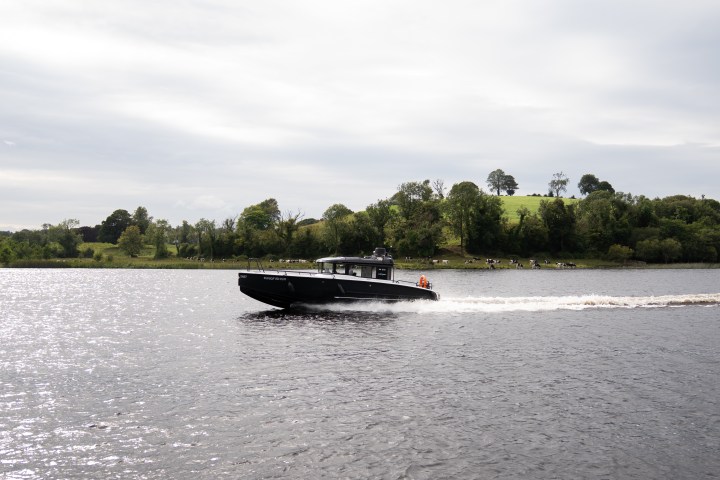 Black motorboat speeding on a river with a green, hilly landscape and trees in the background.