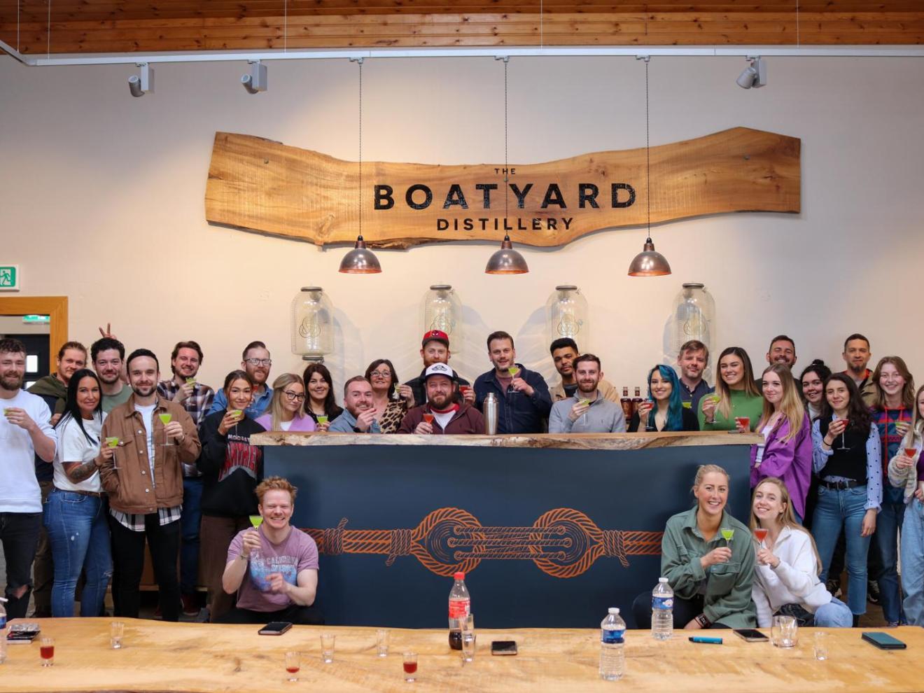 Group of people holding drinks at Boatyard Distillery, standing behind a bar with a wooden sign above.