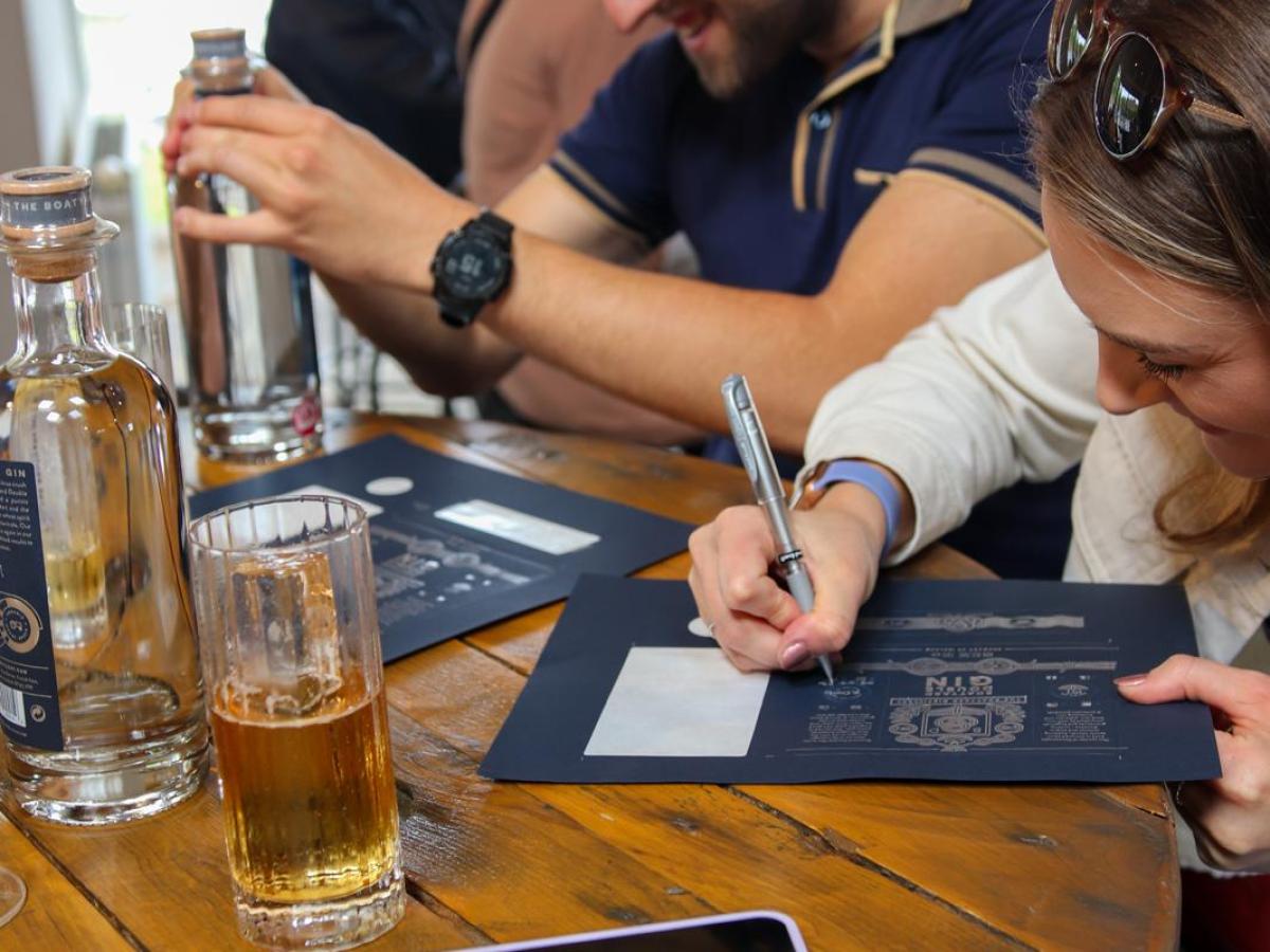 People sitting at a wooden table, writing on papers, with a glass of drink and phone nearby.