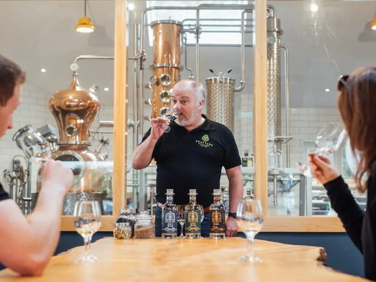 Three people tasting drinks in a distillery with copper equipment in the background.