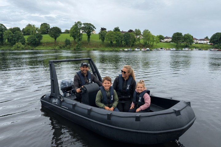 Family of four on a black inflatable boat in a lake with a green shoreline.